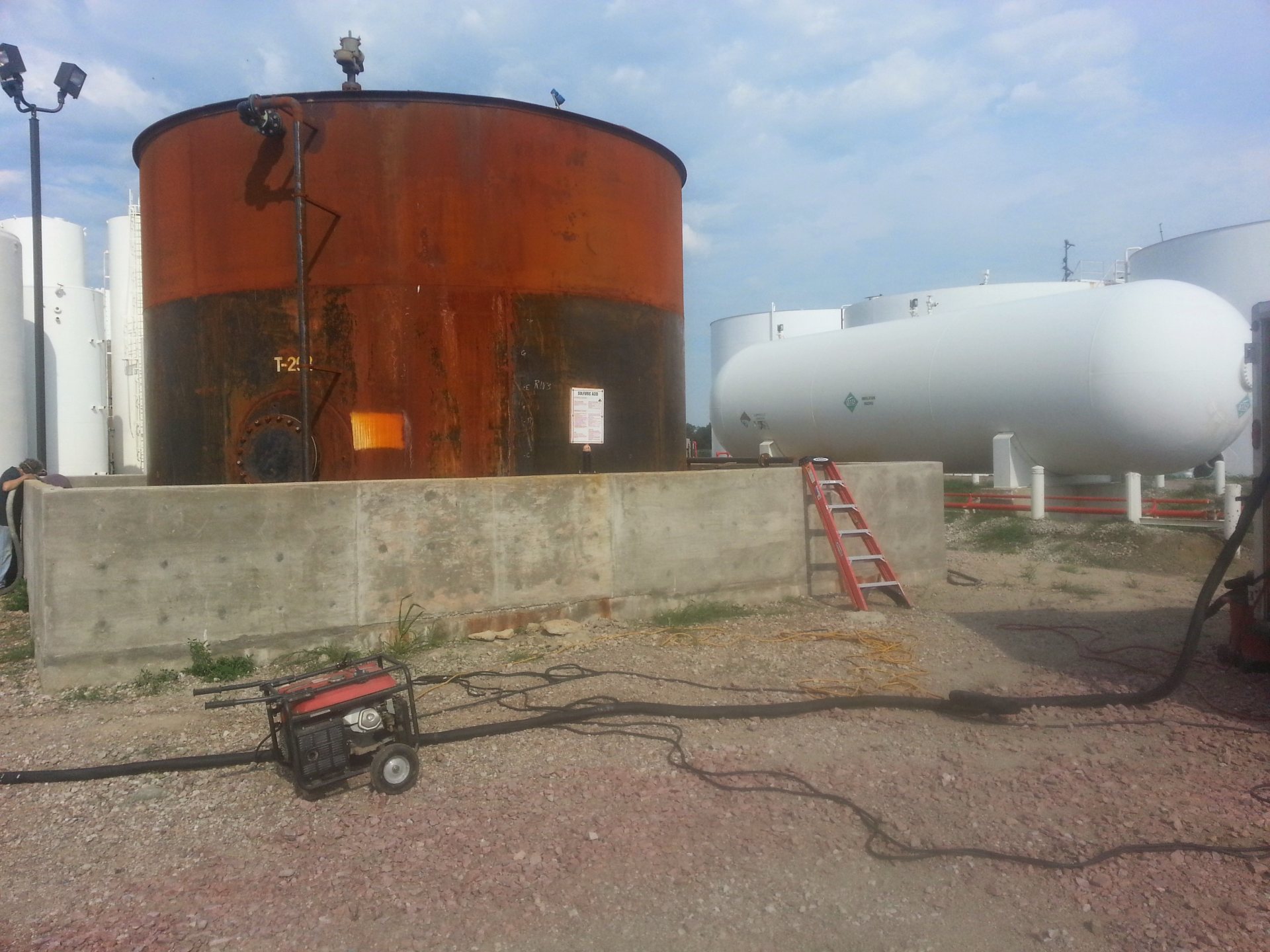 Placarded sulfuric acid storage tank inside a concrete secondary containment dike wall at a tank farm, with coating crew equipment on site